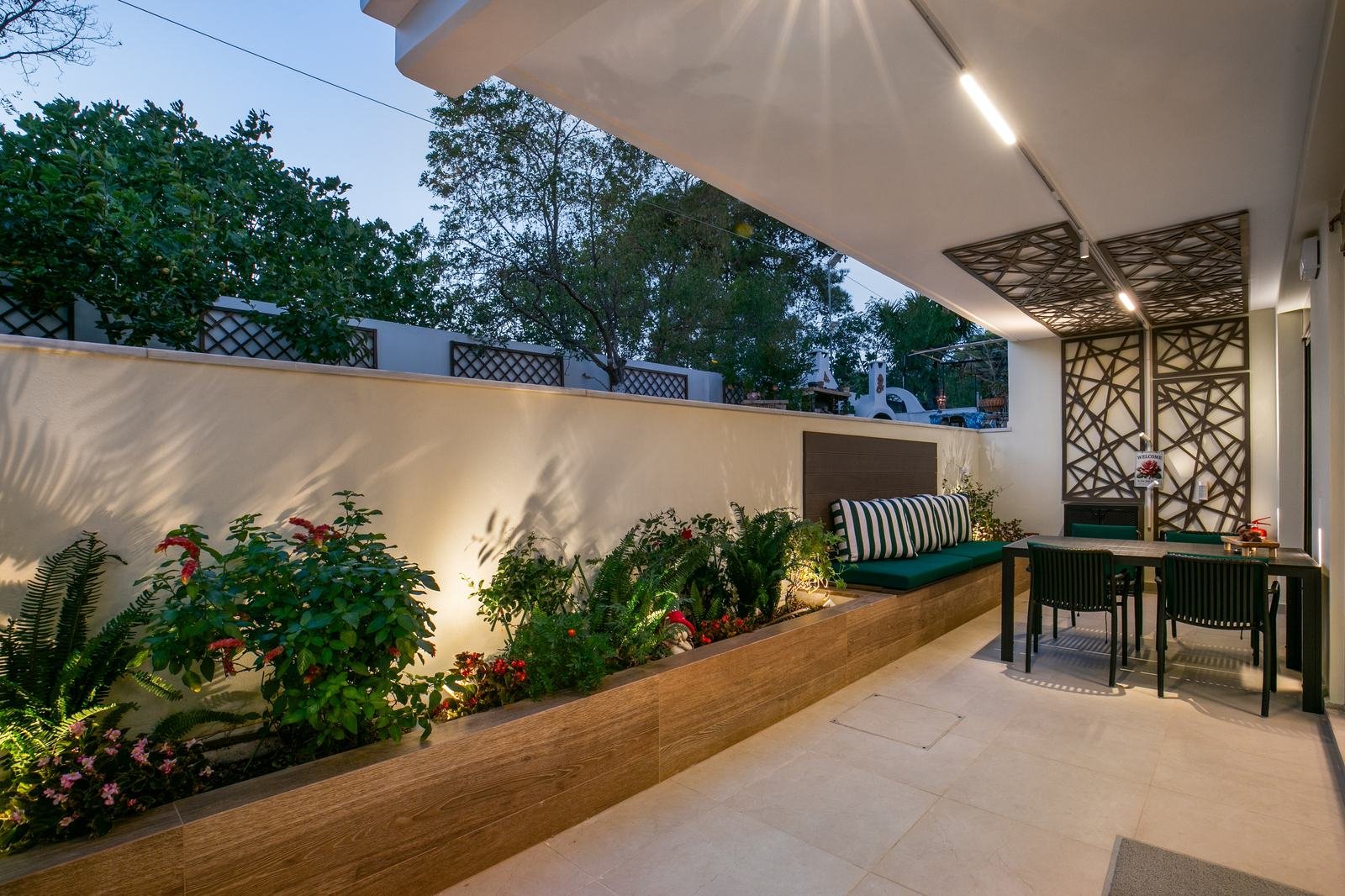 The covered patio of Maria’s Adobe at dusk, with lit planters, a green bench, a dining table and laser-cut pergola panels